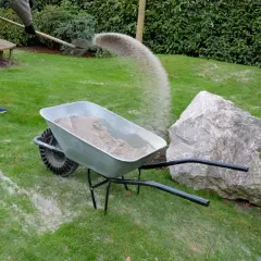Osoba sypie piasek z łopaty do taczki stojącej na trawniku obok kamienia.
