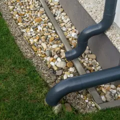 Szare rury spustowe odprowadzają wodę deszczową na kamieniste obrzeże trawnika.