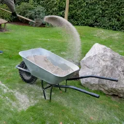 Osoba sypie piasek z łopaty do taczki stojącej na trawniku obok kamienia.
