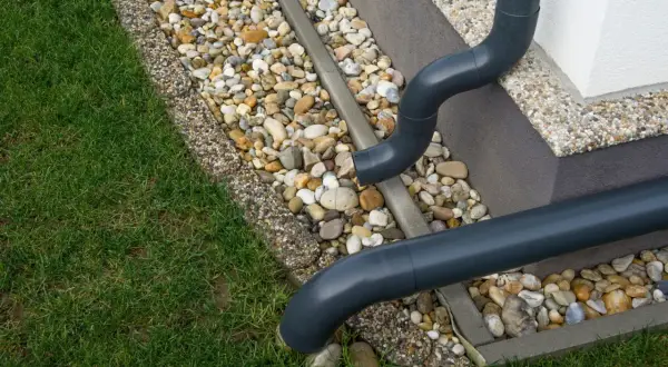 Szare rury spustowe odprowadzają wodę deszczową na kamieniste obrzeże trawnika.