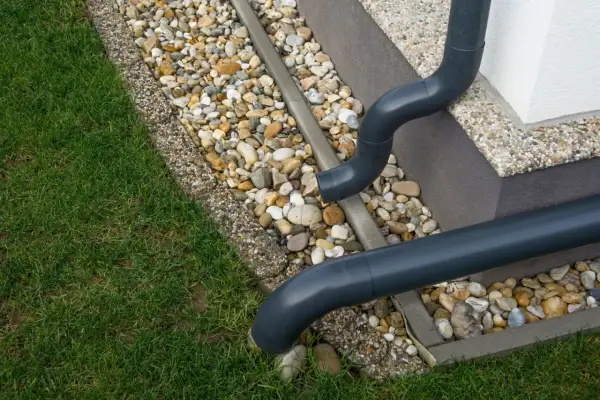 Szare rury spustowe odprowadzają wodę deszczową na kamieniste obrzeże trawnika.